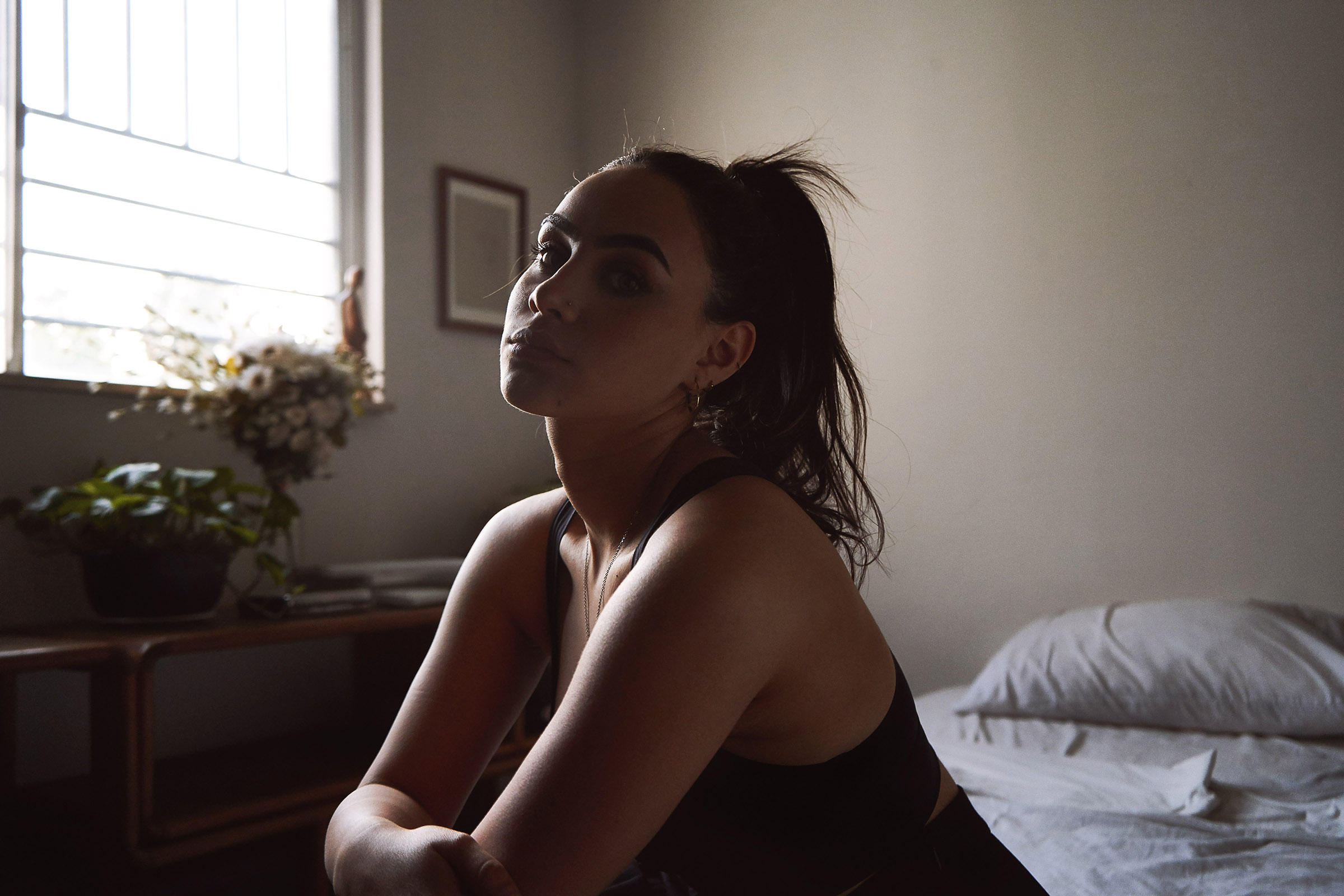Valeria Cisneros a model with a ponytail sits by a window in a room with soft lighting a bed and a table with a plant.